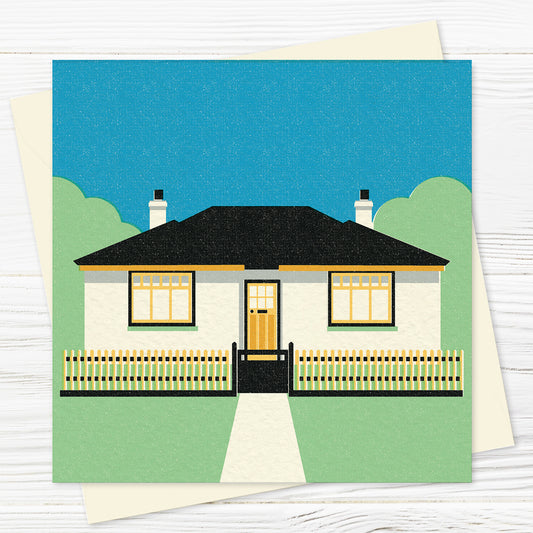 A greetings card and envelope on a wooden table,  featuring a stylised illustration of an art deco bungalow with a picket fence in front in a palette of yellow, black, green and blue