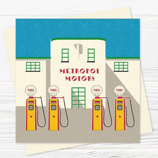 A greetings card and envelope featuring a stylised illustration of a symmetrical, streamlined art deco garage red, green and white against a cloudless blue sky. The words METROPOL MOTORS are written across the front, and there are four red and yellow petrol pumps at the front with the words FUEL on them. 