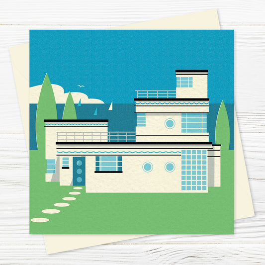 A blank greetings card and envelope  featuring a stylised illustration in green, white and shades of blue of an art deco house by the sea on a sunny day. The house is nautical looking with chrome railings and wavy blue decoration. There are sailing boats on the horizon and white seagulls in the sky. 