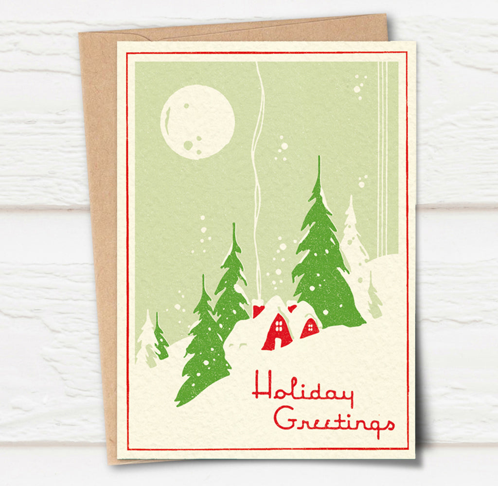 A Christmas card and envelope in red and green featuring a stylised illustration of a cosy house in front of pine trees in a snowy landscape with smoke curling up from the chimney. 