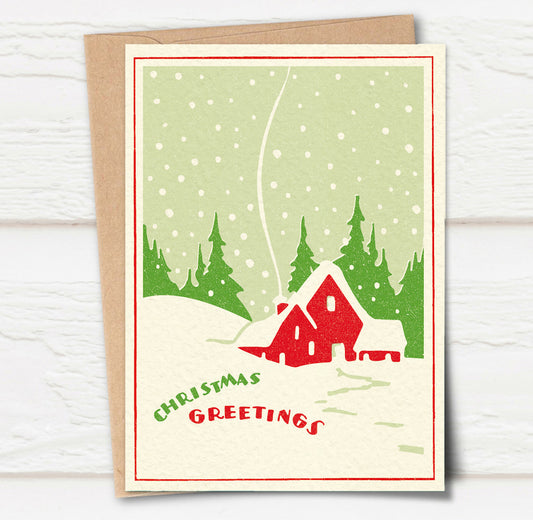 A Christmas card and envelope in red and green featuring a stylised illustration of a cosy house in front of pine trees in a snowy landscape with smoke curling up from the chimney. 