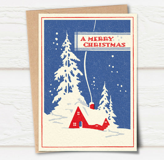 A Christmas card and envelope in red and blue featuring a stylised illustration of a cosy house in front of pine trees in a snowy landscape with smoke curling up from the chimney. 