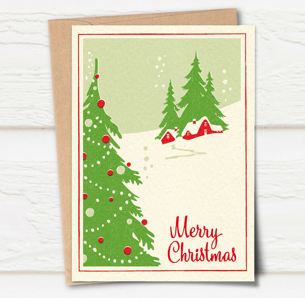 A Christmas card and envelope in red and green featuring a stylised illustration of a cosy house in front of pine trees in a snowy landscape with smoke curling up from the chimney. 