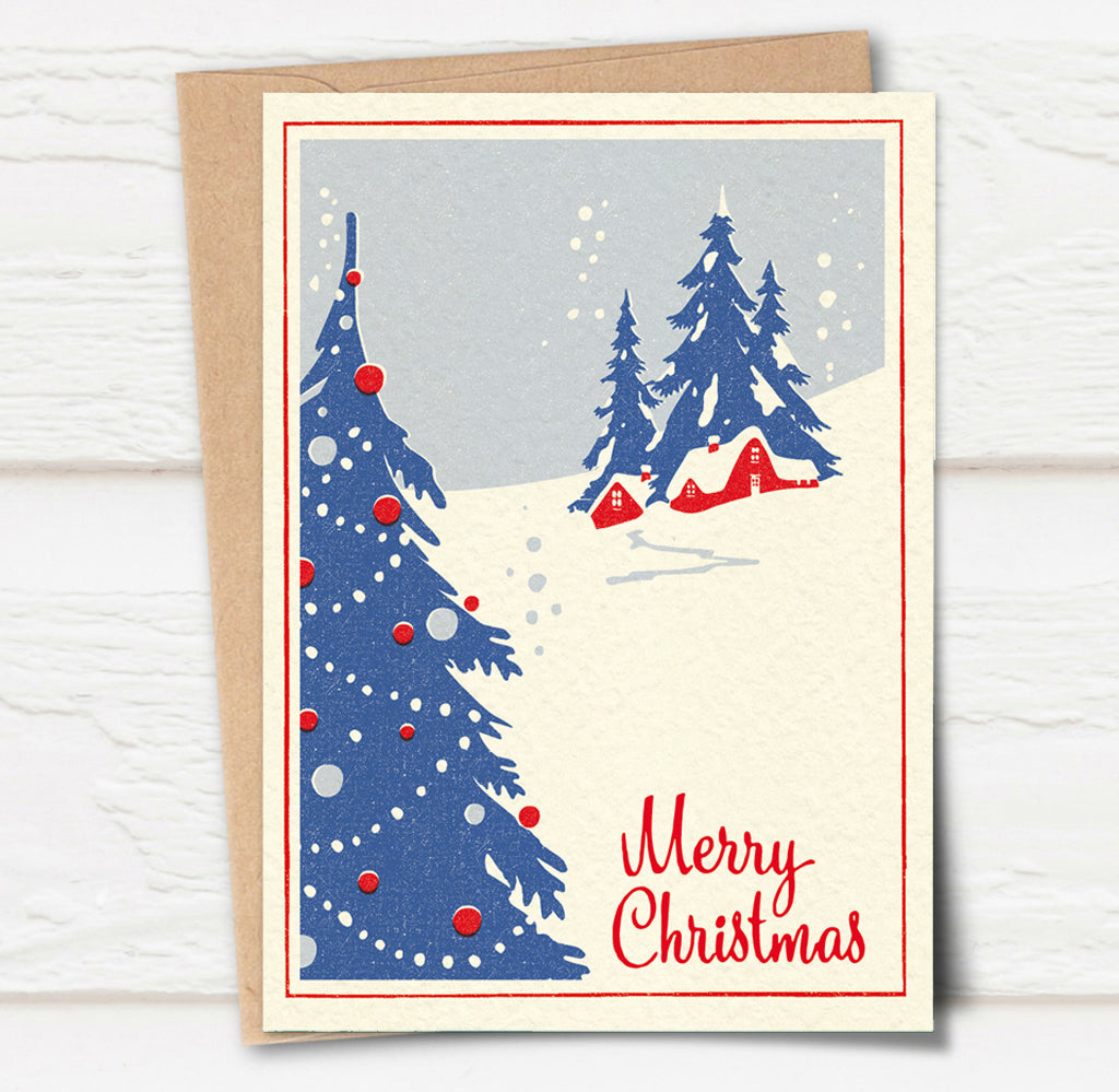 A Christmas card and envelope in red and blue featuring a stylised illustration of a cosy house in front of pine trees in a snowy landscape with smoke curling up from the chimney. 